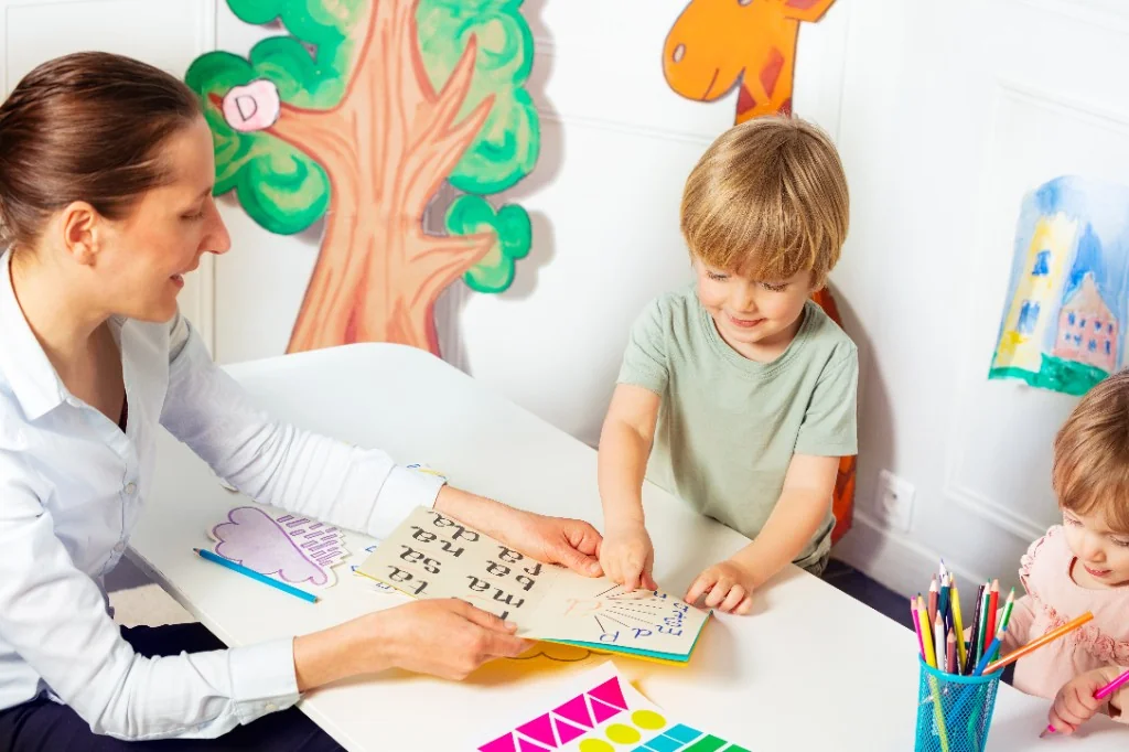 A clinician guides a smiling young child through a letter-matching learning activity at a colorful table in a child-friendly therapy room